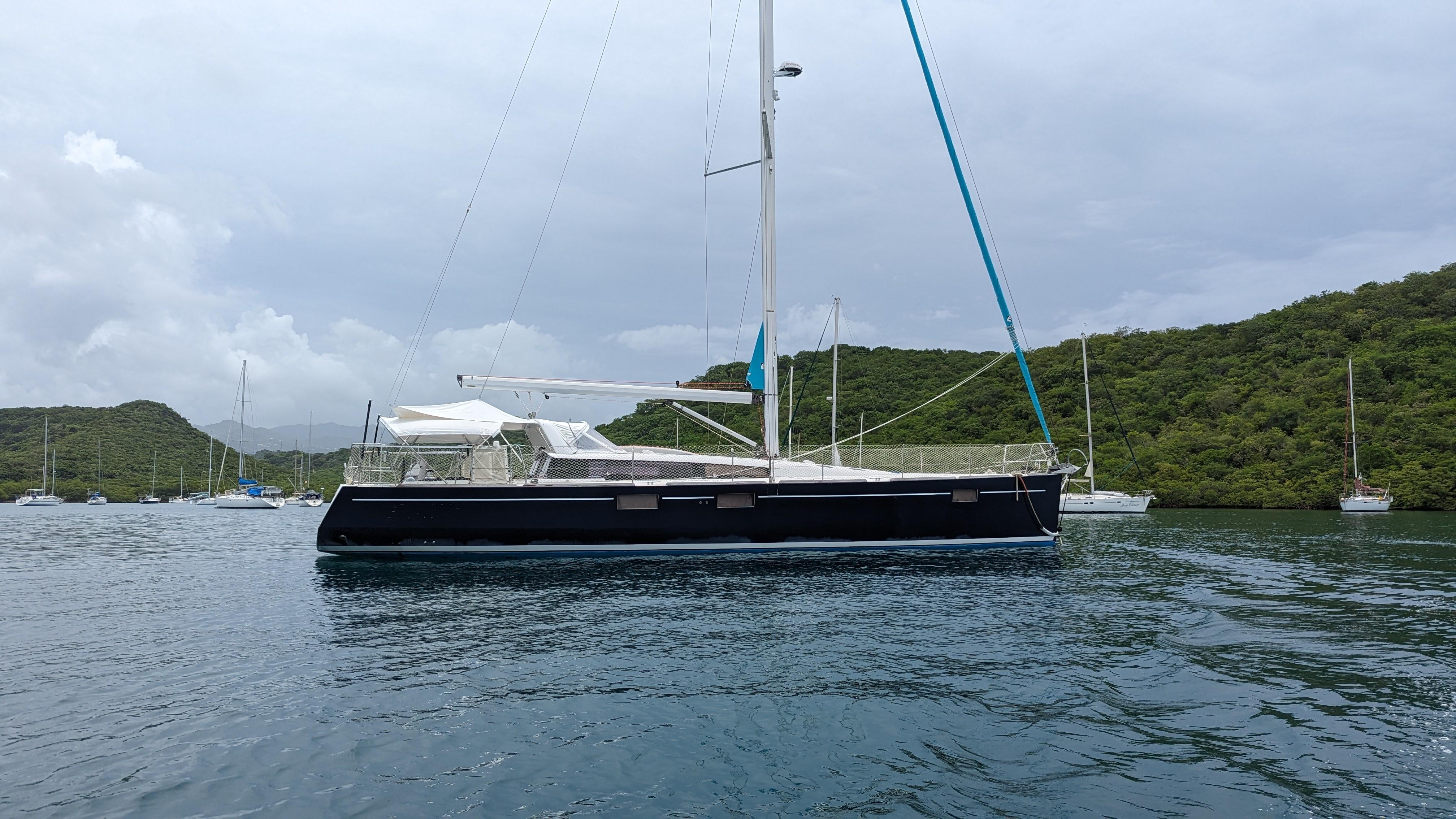 Slide: The Image of 2015 Beneteau Sense 55 sailboat anchored in a scenic bay with lush green hills. - 5