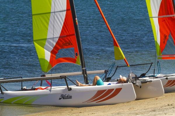 Slide: The Image of 2023 Hobie Cat Getaway sailboats on sandy beach with colorful sails. - 1