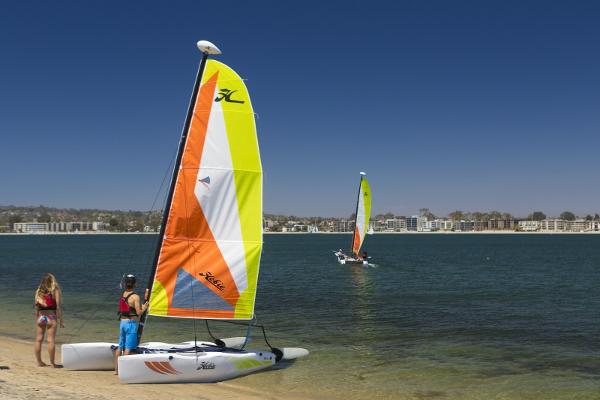 Slide: The Image of 2023 Hobie Cat Wave sailboats on a sunny beach with people preparing to sail. - 1