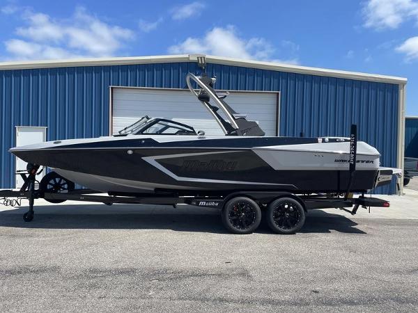 Slide: The Image of 2023 Malibu 24 MXZ boat on trailer in front of blue warehouse. - 0