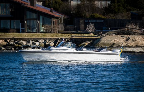 Slide: The Image of 2025 Nimbus T9 boat cruising near a rocky shoreline with houses in the background. - 0