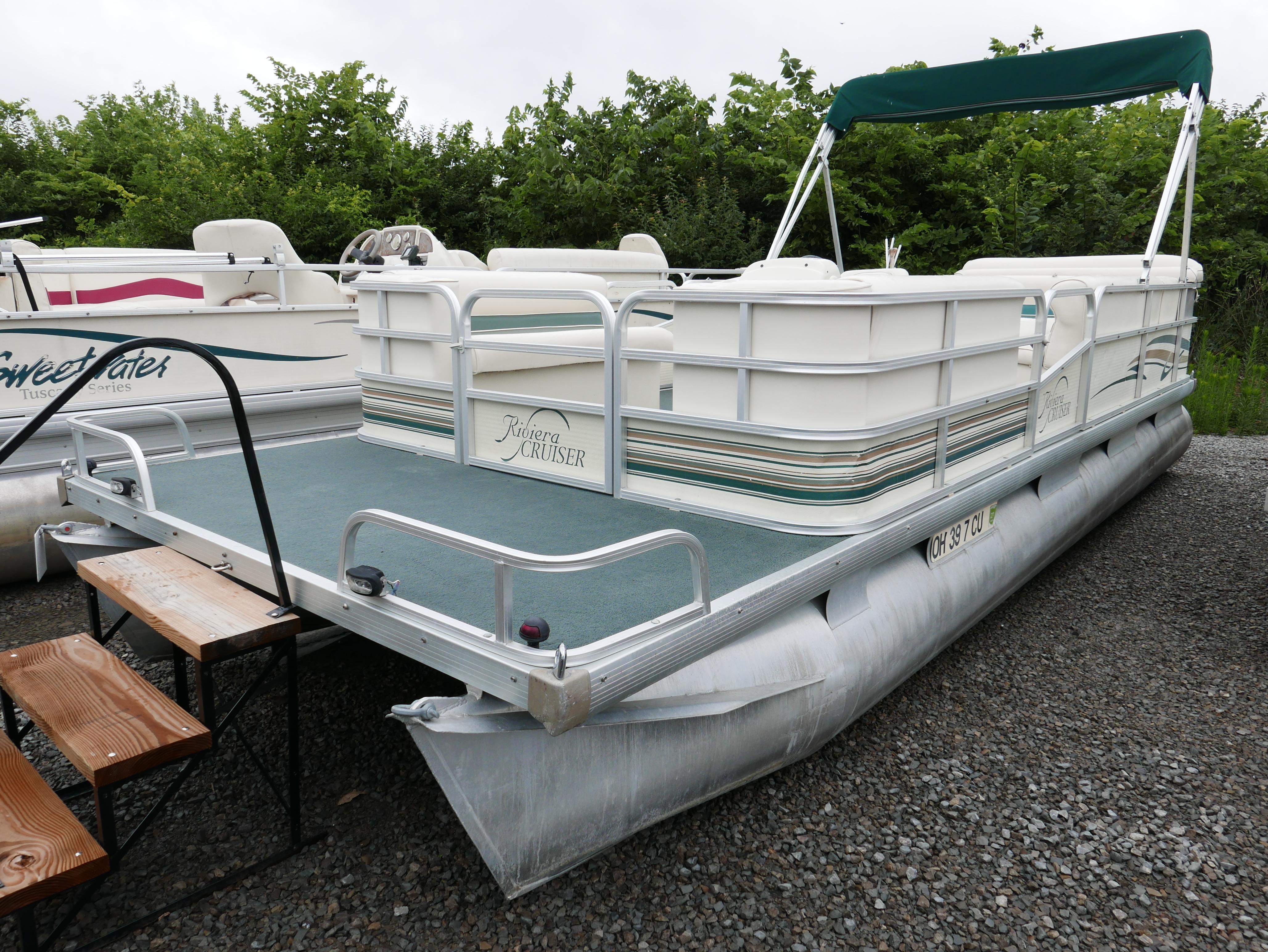 Slide: The Image of 2000 Riviera Cruiser pontoon boat with green canopy, parked on gravel. - 2