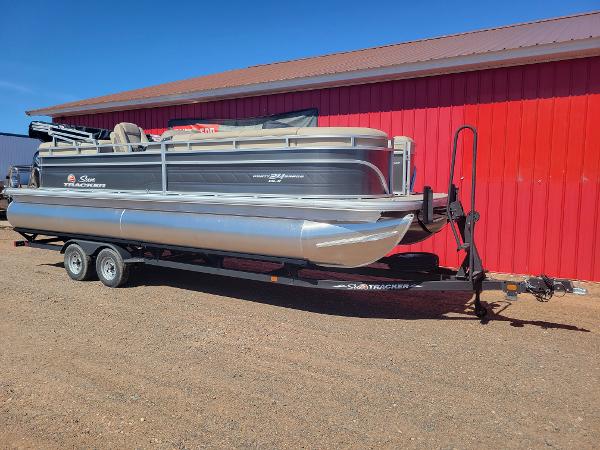 Slide: The Image of 2024 Sun Tracker Party Barge 24 DLX pontoon boat on trailer, parked near red building. - 1