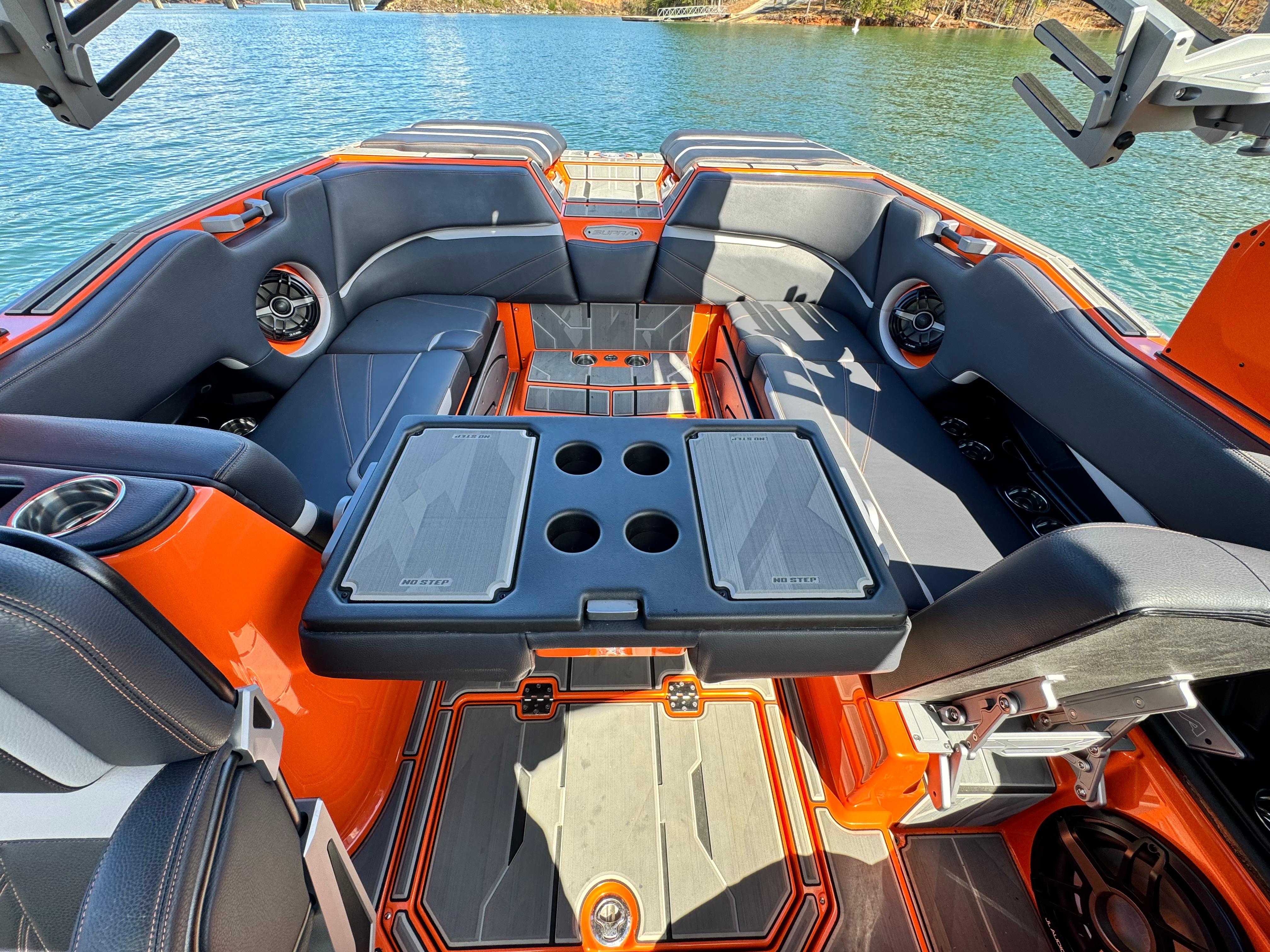 Slide: The Image of 2022 Supra SL 550 boat interior with orange and gray seating, table, and cup holders on a lake. - 7