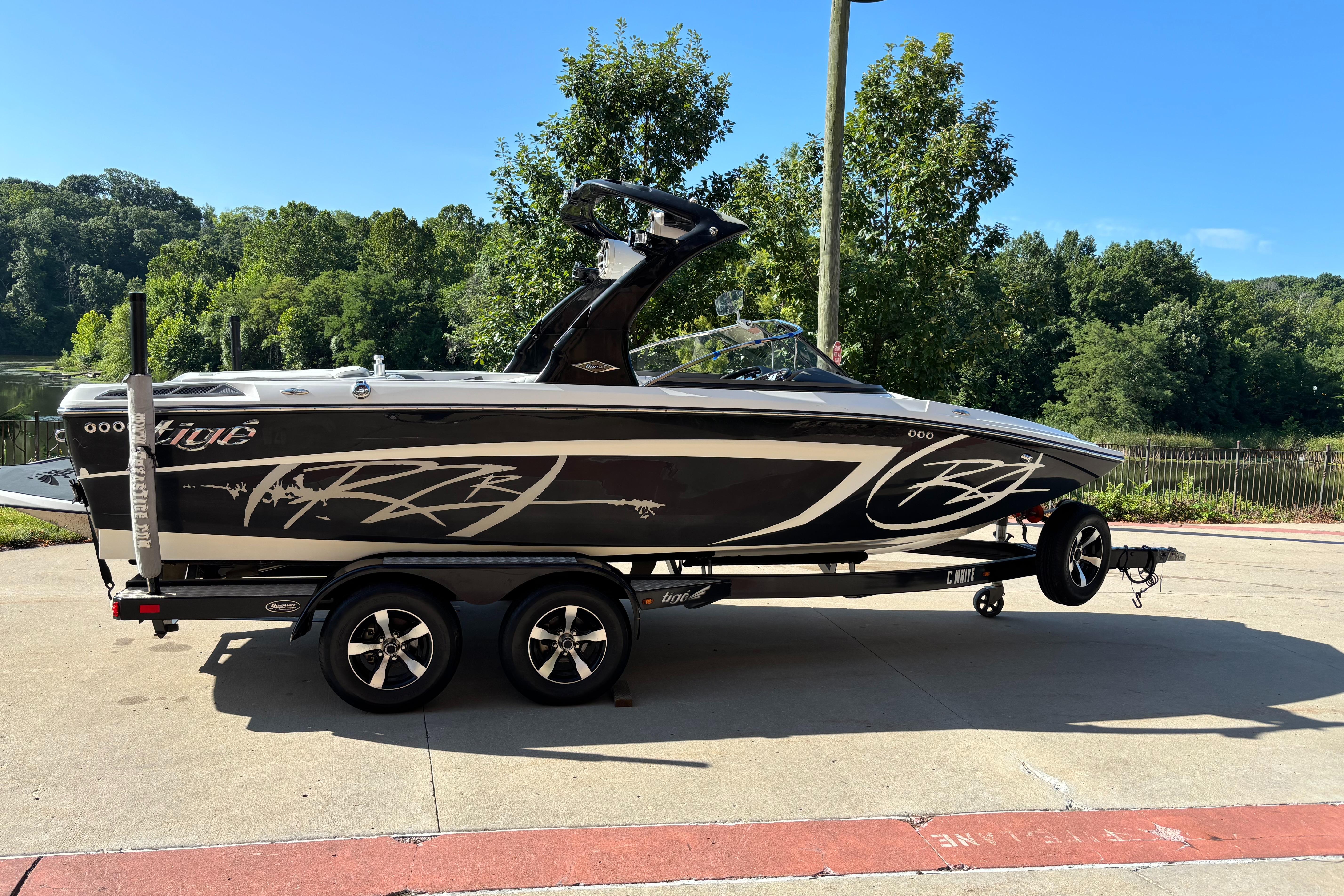 Slide: The Image of 2014 Tigé RZR boat on trailer, parked near a scenic lake with lush greenery. - 0